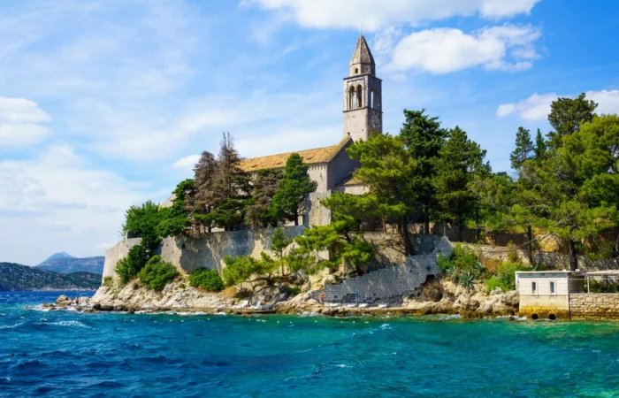 Historic Franciscan monastery and bell tower surrounded by pine trees on Lopud Island, Croatia, with stone walls rising above the blue Adriatic Sea.