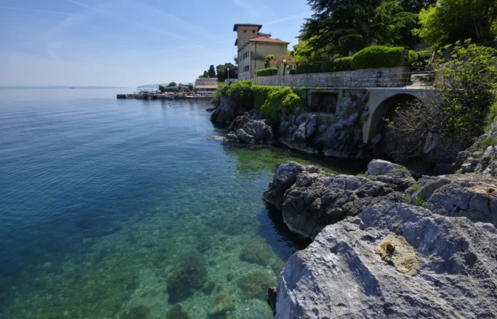 Elegant historic villa above rocky coastline and Lungomare promenade in Lovran, Croatia, with crystal-clear blue Adriatic waters and lush Mediterranean greenery.
