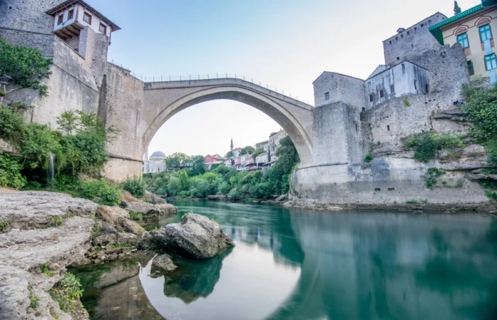 Stone arch of Stari Most (Old Bridge) in Mostar, Bosnia and Herzegovina, viewed from the riverbank with Ottoman towers and historic old town reflected in the calm Neretva River.