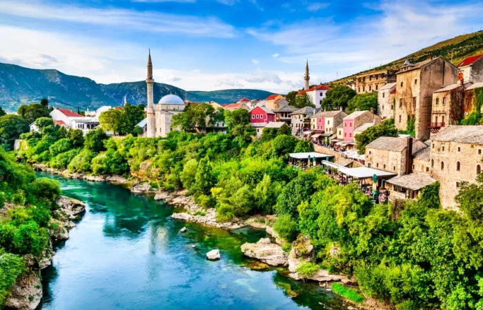 Panoramic view of Mostar Old Town with mosque minarets, colorful Ottoman houses, and the emerald Neretva River flowing through lush greenery in Bosnia and Herzegovina.