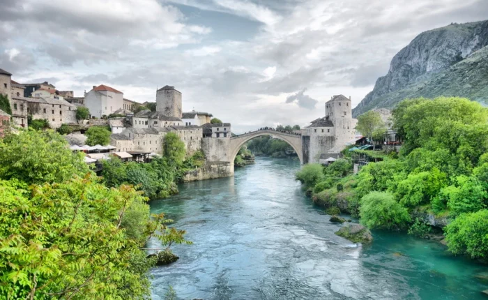 Wide-angle view of Mostar’s Stari Most (Old Bridge) with Ottoman towers, stone houses, and lush greenery spanning the emerald Neretva River, Bosnia and Herzegovina.