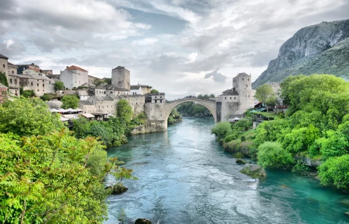 Wide-angle view of Mostar’s Stari Most (Old Bridge) with Ottoman towers, stone houses, and lush greenery spanning the emerald Neretva River, Bosnia and Herzegovina.