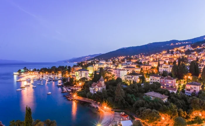 Aerial panoramic twilight view of Opatija Riviera, Croatia, with illuminated city, harbor, boats, and sloping green hills along the Adriatic coast.