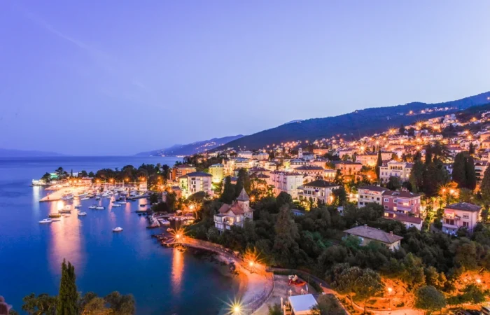 Aerial panoramic twilight view of Opatija Riviera, Croatia, with illuminated city, harbor, boats, and sloping green hills along the Adriatic coast.