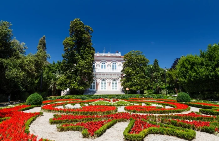 Villa Angiolina surrounded by colorful flower beds and lush gardens in Opatija, Croatia, beneath a deep blue sky.