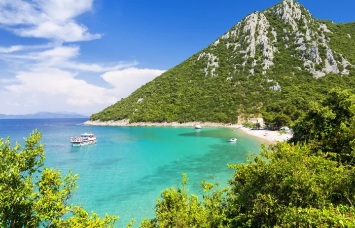 Panoramic view of Divna Beach on the Pelješac peninsula, Croatia, featuring turquoise sea, sandy-pebble shoreline, green hills, and anchored boats in a sheltered bay.