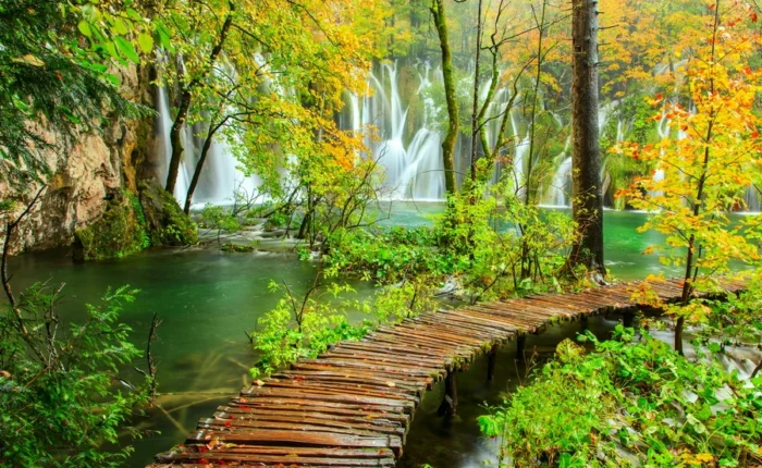Wooden boardwalk in autumn with golden leaves and waterfalls in Plitvice Lakes National Park, Croatia, surrounded by forest and emerald water.