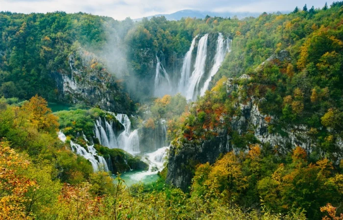 Panoramic autumn view of Plitvice Lakes waterfalls pouring over cliffs into a misty, golden forest in Croatia.