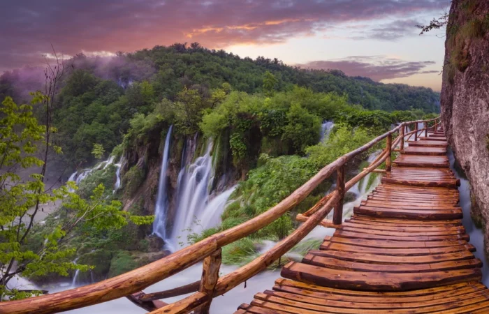 Wooden boardwalk with railings stretches above stunning waterfalls at sunset in Plitvice Lakes National Park, Croatia.