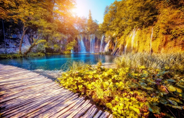 Wooden boardwalk curving along a turquoise lake at sunrise, with golden light illuminating waterfalls and lush foliage in Plitvice Lakes National Park, Croatia.