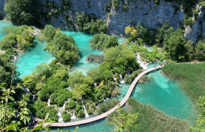 Aerial view of a curved wooden boardwalk crossing crystal-clear turquoise pools and islands at Plitvice Lakes National Park, Croatia.