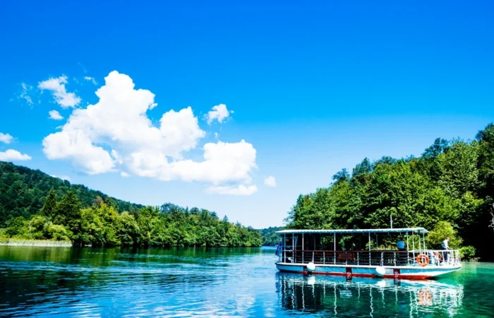 Electric boat sailing on the crystal-clear waters of Plitvice Lakes National Park, Croatia, with green forests reflected under a bright sky.