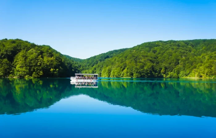 Electric sightseeing boat gliding across Kozjak Lake in Plitvice Lakes National Park, Croatia, with perfect reflections of green hills and blue sky.