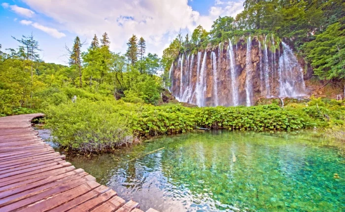 Wide view of Veliki Slap-the Great Waterfall-cascading into a turquoise lake at Plitvice Lakes National Park, Croatia, with lush forest and wooden walking path.