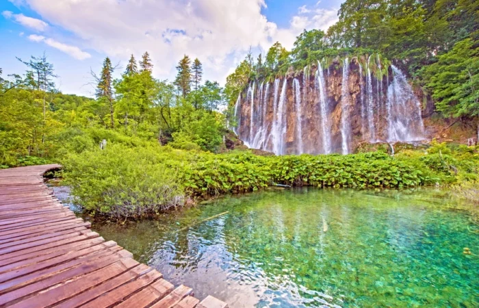 Wide view of Veliki Slap-the Great Waterfall-cascading into a turquoise lake at Plitvice Lakes National Park, Croatia, with lush forest and wooden walking path.