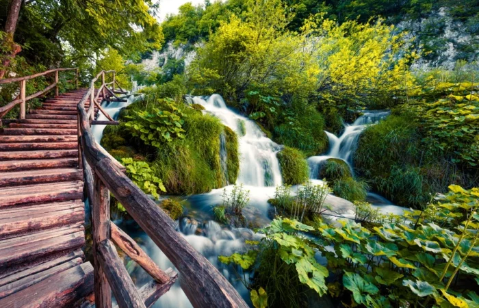 Lush green waterfalls and a rustic wooden trail surrounded by vibrant spring vegetation in Plitvice Lakes National Park, Croatia.