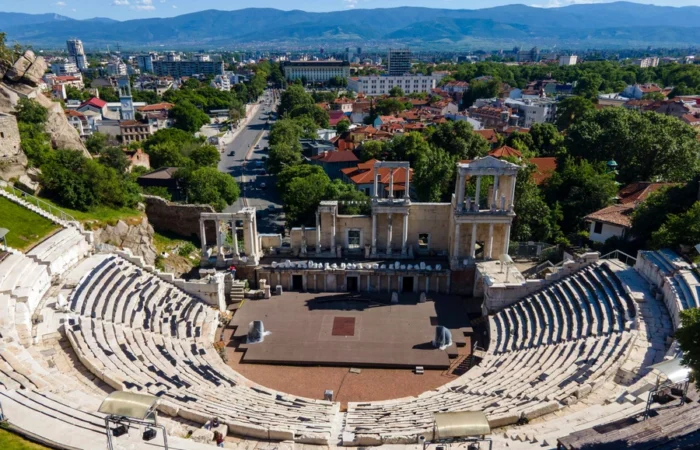 Aerial view of Plovdiv’s Ancient Roman Theatre with marble seating, stage, and panoramic cityscape backed by mountains in Bulgaria.