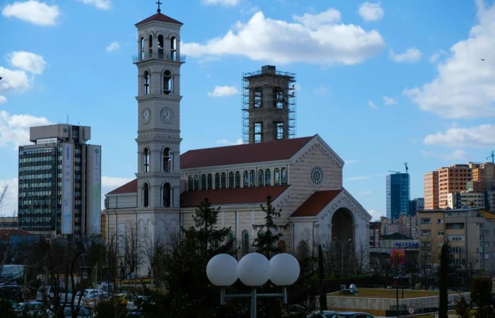 Cathedral of Saint Mother Teresa in Pristina with white bell towers, red-tiled roof, and city skyline under a blue, partly cloudy sky.