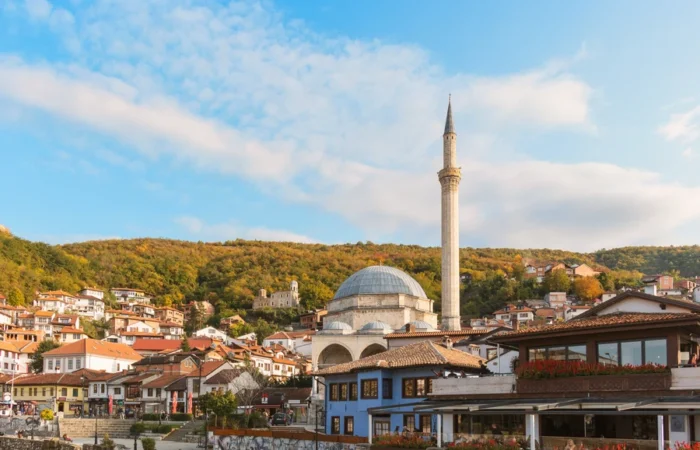 Sinan Pasha Mosque rises above Prizren’s red-roofed old town, set against tree-covered hills and a vivid blue sky in Kosovo.