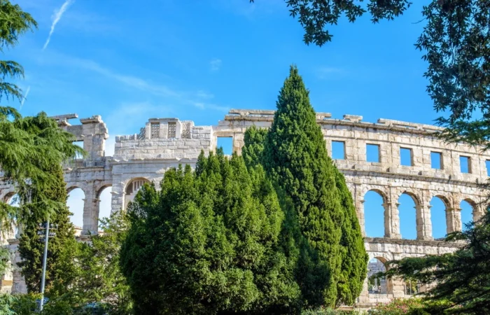 Roman stone arches of Pula Arena framed by lush gardens and tall trees on a bright day in Istria, Croatia.