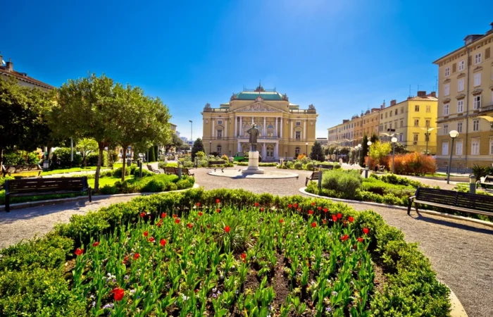 Ivan Zajc Croatian National Theatre in Rijeka, Croatia, viewed from a landscaped park with spring flower beds and benches under a clear blue sky.