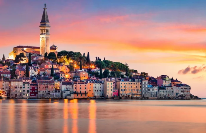 Colorful houses and the bell tower of St. Euphemia glow against a pastel sky and calm sea at sunset in Rovinj, Croatia.