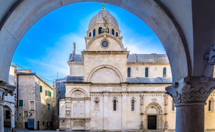 The white stone Cathedral of St. James in Šibenik, Croatia, framed by an arch in the historic old town on a clear blue day.