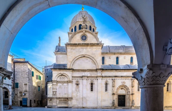 The white stone Cathedral of St. James in Šibenik, Croatia, framed by an arch in the historic old town on a clear blue day.