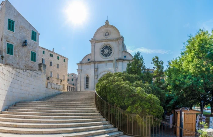 Sunny day view of Šibenik Cathedral’s iconic facade and rose window, seen from stone staircase and medieval old town houses.