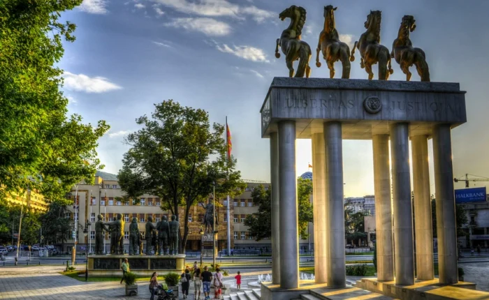 Sunset over Skopje’s Porta Macedonia monument with four bronze horses atop neoclassical columns and a nearby group sculpture, framed by city greenery.