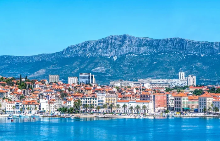 Bright cityscape of Split, Croatia, with the waterfront Riva promenade, Adriatic harbor, and the forested slopes of Marjan mountain in the background.
