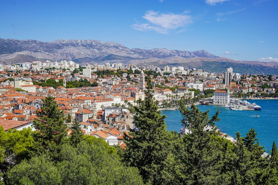 View across the green slopes of Marjan hill to the city of Split, with the red-tiled Old Town, Riva promenade, blue Adriatic, and city harbor.