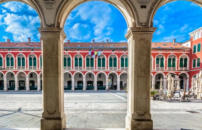 Neoclassical arcades and red facades frame Republic Square (Prokurative), Split, Croatia, seen through stone arches under a blue sky.