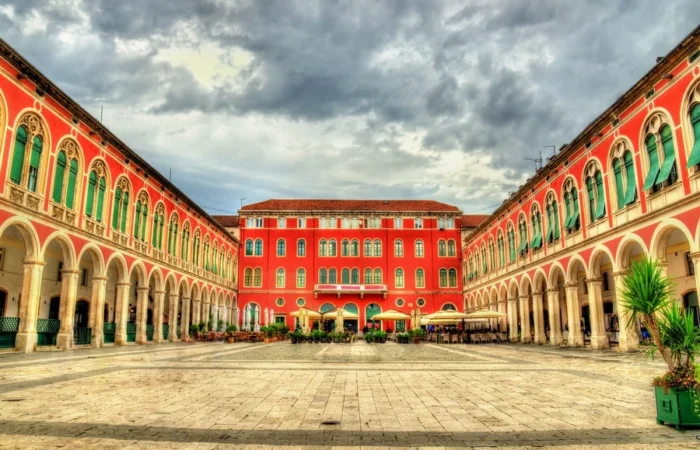 The red arcades and neoclassical facades of Republic Square (Prokurative) in Split, Croatia, under dramatic clouds.