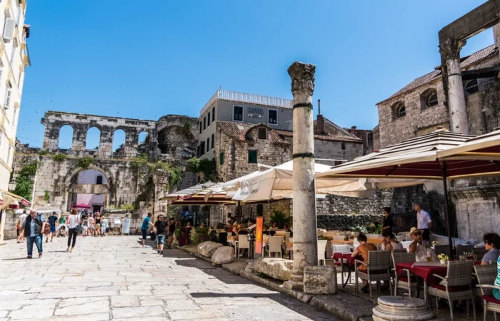 Outdoor café tables sit among ancient Roman columns and ruins by Split’s Silver Gate in Diocletian’s Palace, Croatia, on a sunny day.