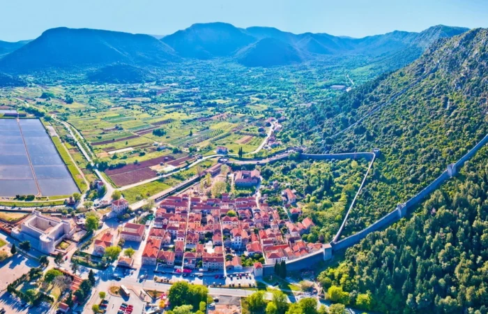Aerial view of Ston, Croatia, revealing medieval stone city walls stretching over forested hills, red-roofed town center, and historic salt pans on the Pelješac peninsula.