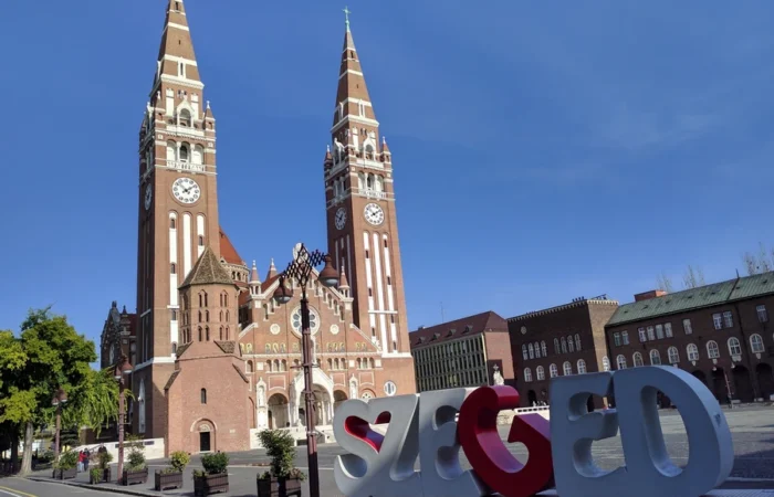 The twin spires of Szeged Votive Church rise above Dóm Square, with a modern Szeged sign and clear blue sky in Hungary’s vibrant university city.