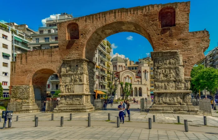 The Arch of Galerius, a Roman-era triumphal arch in central Thessaloniki, Greece, with historic reliefs surrounded by city life and modern buildings.
