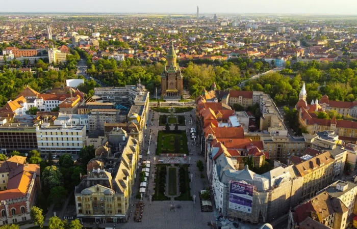 Aerial view of Timișoara’s Victory Square with the Romanian Orthodox Metropolitan Cathedral, surrounded by historic buildings and lush green cityscape.