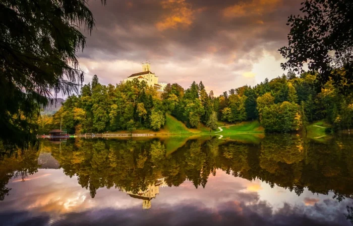 Trakošćan Castle crowns a hill above a forest ablaze with autumn colors, perfectly reflected in the still lake at sunset, northern Croatia.