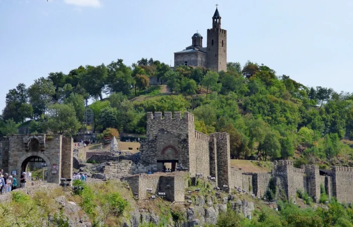 The ancient stone walls of Tsarevets Fortress crown a lush green hill in Veliko Tarnovo, Bulgaria, with the Patriarchal Cathedral visible atop the summit.