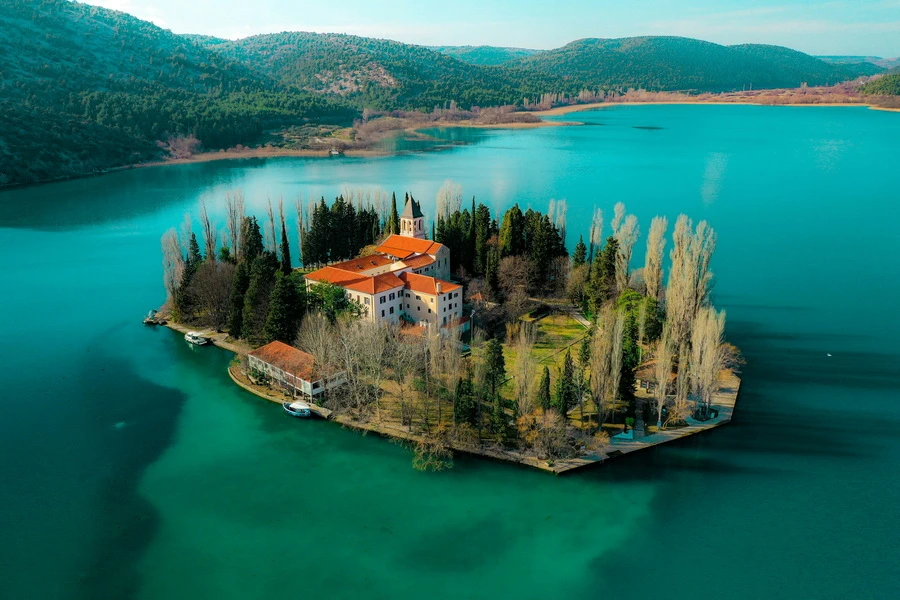 Aerial view of the historic monastery and lush trees on Visovac Island in turquoise Lake Krka, surrounded by green hills in Croatia.