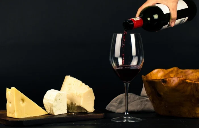 Red wine is poured into a glass next to an artisanal cheese assortment on a dark background, conveying a gourmet wine and food pairing experience.
