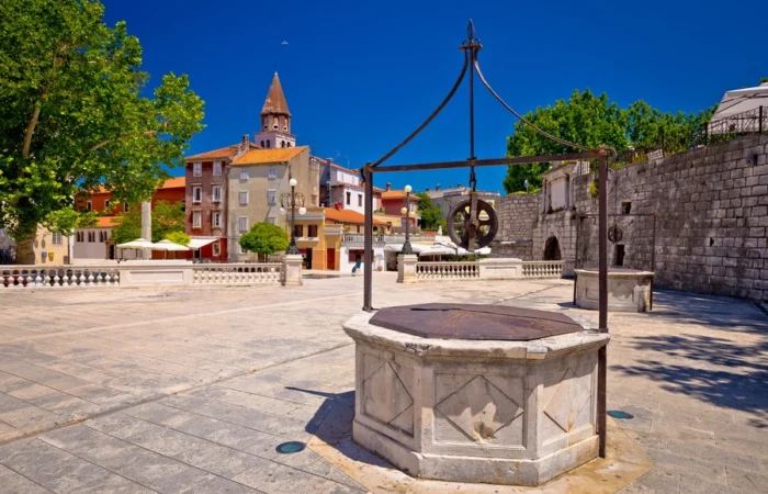 Five ornate historic stone wells line Trg pet bunara (Five Wells Square) in Zadar, Croatia, with old city walls and the Captain’s Tower seen under a clear sky.