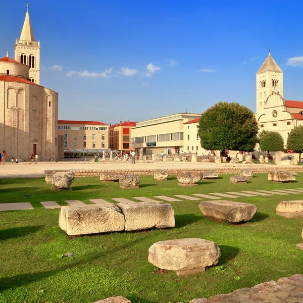 Stones of the Roman Forum lie in front of St. Donatus Church and the Cathedral of St. Anastasia in Zadar’s old town, under a blue sky.