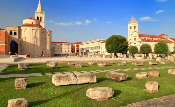 Stones of the Roman Forum lie in front of St. Donatus Church and the Cathedral of St. Anastasia in Zadar’s old town, under a blue sky.