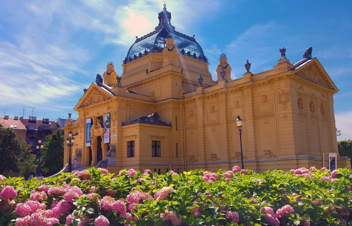 Zagreb’s Art Pavilion with a majestic blue dome, ornate yellow facade, and lush hydrangea blooms in the foreground under a bright summer sky.