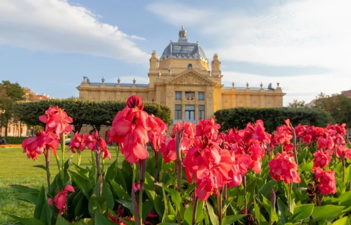 Close-up of red canna lilies in front of Zagreb’s yellow Art Pavilion on a sunny summer day in Croatia, with blue sky and lush gardens.