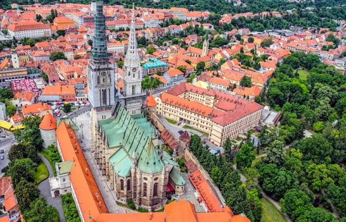 Spectacular aerial view of Zagreb Cathedral’s twin towers, green copper roof, and the red-tiled historic rooftops of Croatia’s capital city, set amid lush parks and gardens.