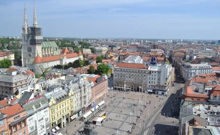 Panoramic aerial photo of Zagreb’s Ban Jelačić Square, the city’s bustling central plaza, colorful historic buildings, tram tracks, and Zagreb Cathedral’s twin spires in the distance.